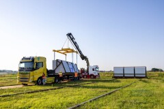An electric truck delivering a coated flood barrier panel for the Arcen dike project, supported by a crane and construction crew.