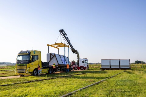An electric truck delivering a coated flood barrier panel for the Arcen dike project, supported by a crane and construction crew.